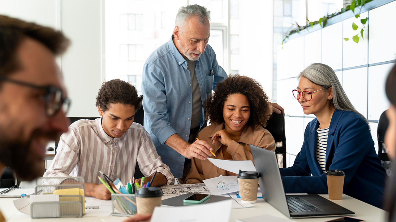 A group of business professionals are gathered around a table in a bright, clean workspace. A man stands over a younger woman to review her work.