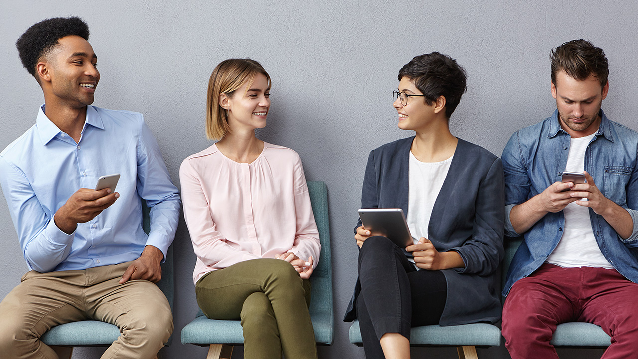 A group of young professionals sit in chairs lined against a wall. Several smile at each other, some have phones or tablets.